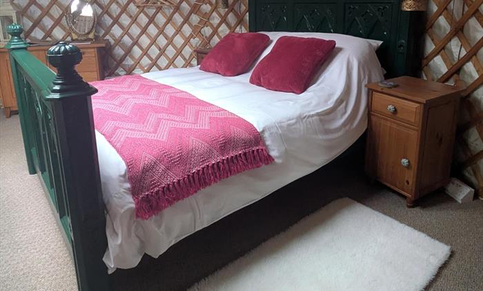An ornate, tall, Gothic style, green, king-size bed . It has white bedding, a pink blanket and deep pink velvet cushions. There are wood bedside cabin